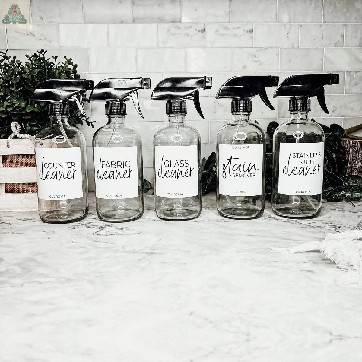 Five 16oz Glass Cleaning Bottles labeled for counter, fabric, glass, stain remover, and stainless steel cleaners are arranged in a row on a marble countertop with green plants in the background.
