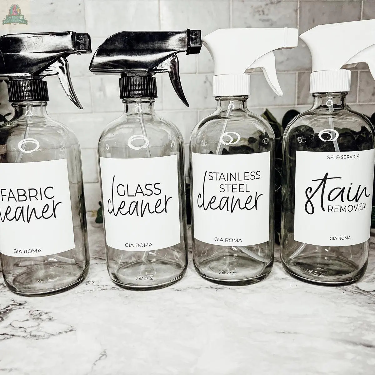 Four 16oz Glass Cleaning Bottles with black and white nozzles are labeled Fabric Cleaner, Glass Cleaner, Stainless Steel Cleaner, and Stain Remover. They’re arranged on a marble countertop against a tiled backsplash.