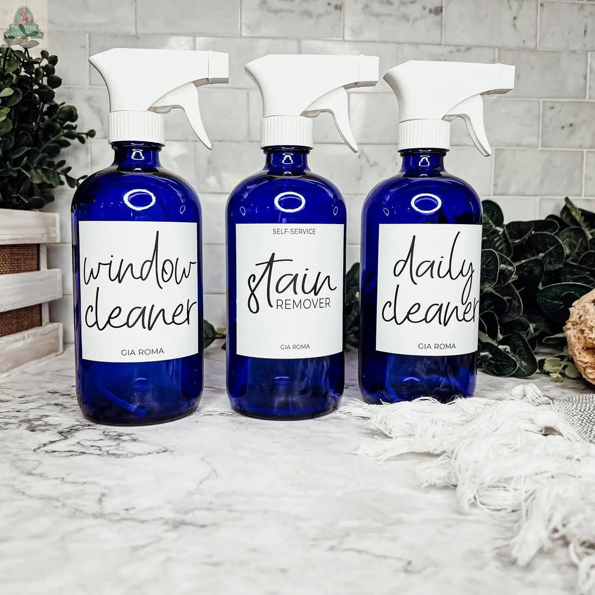 Three 16oz Glass Cleaning Bottles labeled Window Cleaner, Stain Remover, and Daily Cleaner are displayed on a marble countertop with greenery and a white towel in the background.