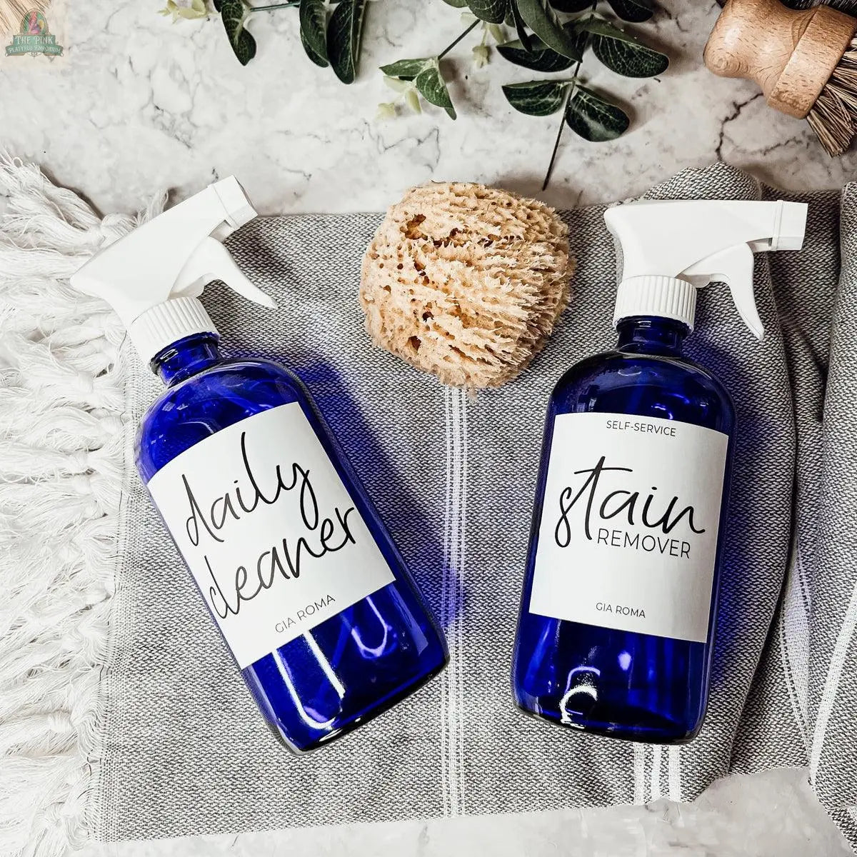 Two blue 16oz Glass Cleaning Bottles labeled Daily Cleaner and Stain Remover rest on a gray striped cloth beside a natural sponge and greenery, all arranged on a marble surface.