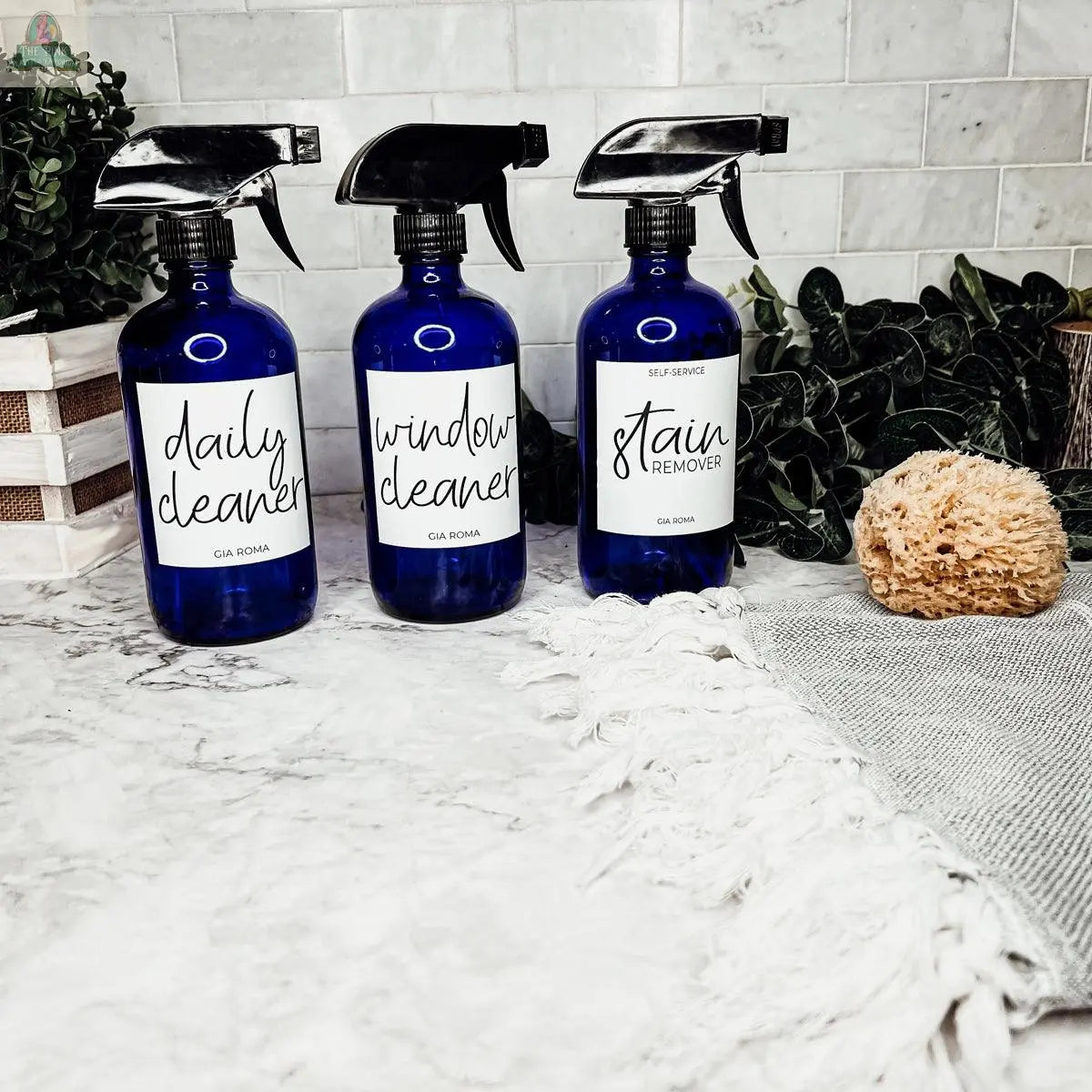 Three blue 16oz Glass Cleaning Bottles—labeled daily cleaner, window cleaner, and stain remover—are displayed on a marble countertop with a sponge, textured towel, and green plants in the background.