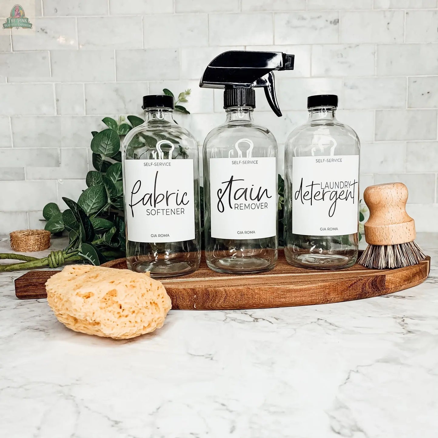 Three 16oz Glass Clear bottles with white labels—fabric softener, stain remover, and detergent—are arranged on a wooden tray next to a sponge, scrub brush, and green leaves on a marble countertop with a white tiled backsplash.
