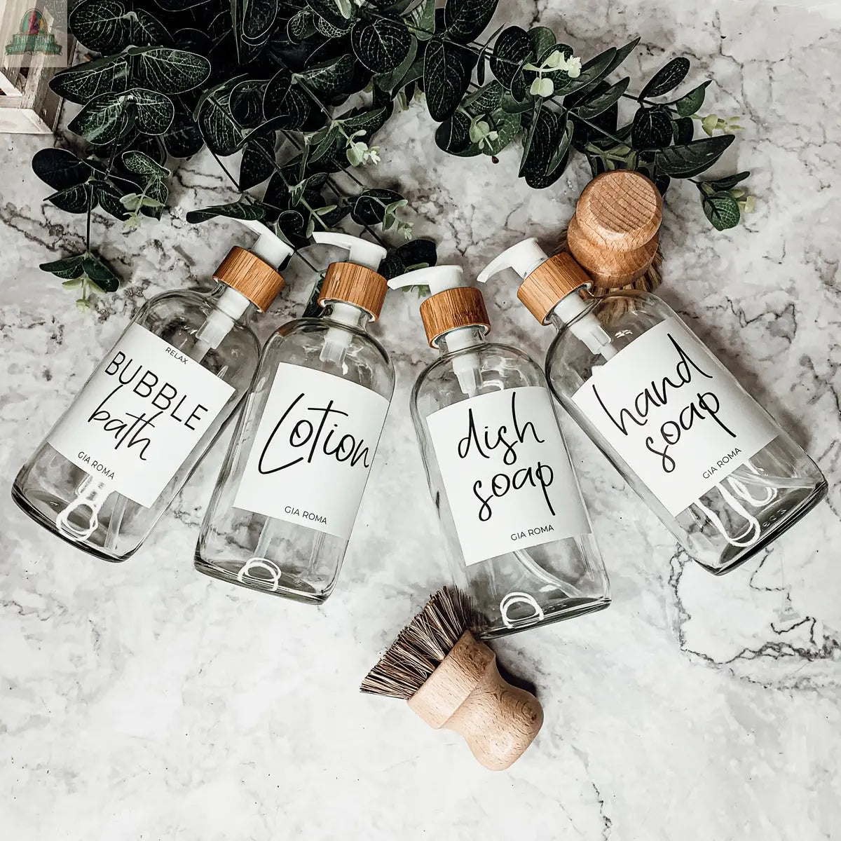 Four 16oz Glass Clear | White Label dispenser bottles—labeled Bubble Bath, Lotion, Dish Soap, and Hand Soap—sit on a marble surface with a wooden scrub brush and greenery in the background.