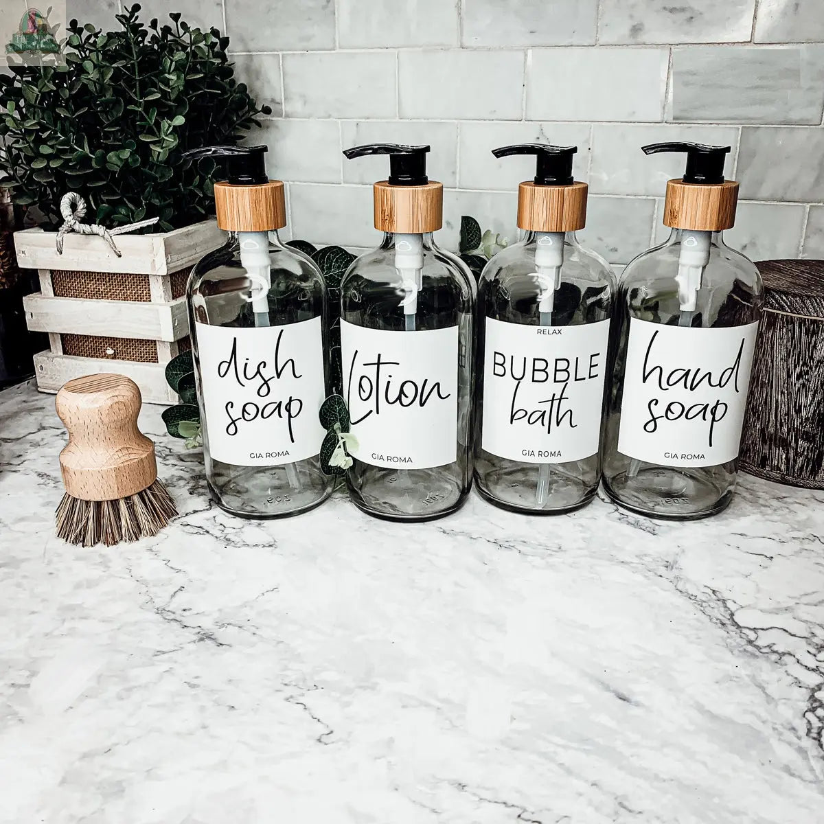 Four 16oz Glass Clear | White Label dispenser bottles for dish soap, lotion, bubble bath, and hand soap sit on a marble counter with a wooden scrub brush and basket of towels, set against a tiled backsplash.