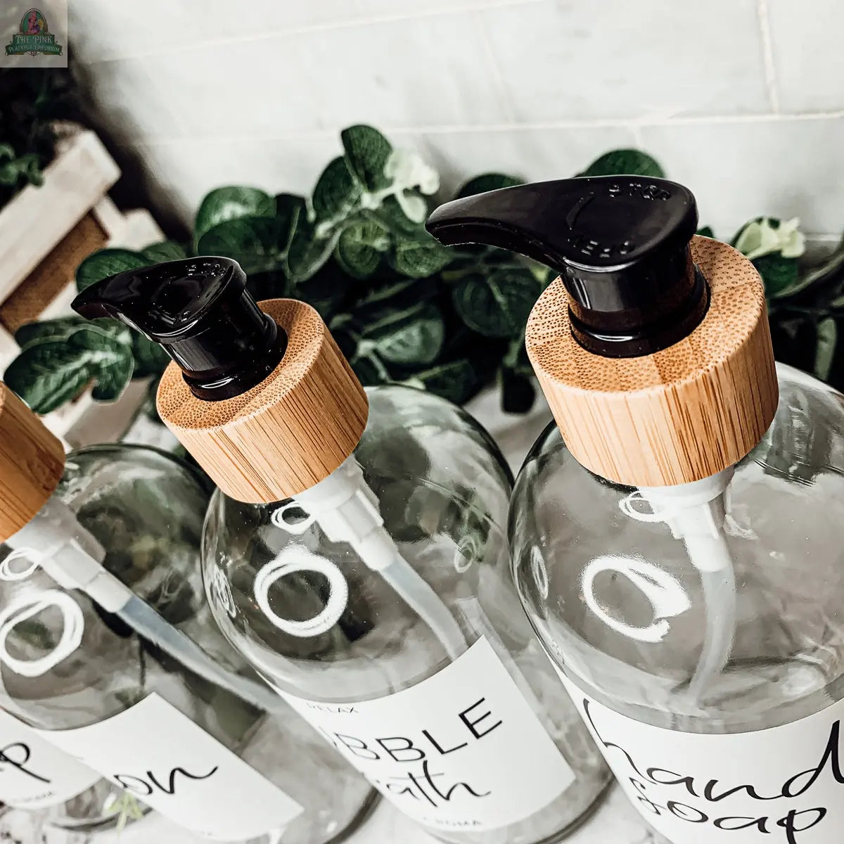 Three 16oz Glass Clear dispensers with bamboo lids and black pumps, each featuring a white waterproof label for “Hand Soap,” “Bubble Bath,” or “Lotion,” are displayed on a white surface with green plants in the background.