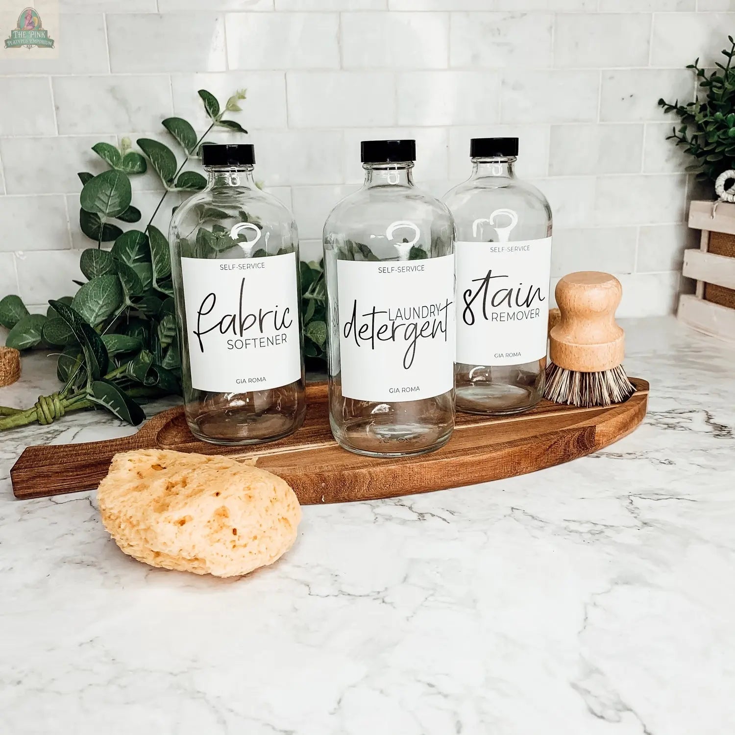 Three 16oz Glass Clear bottles with white waterproof labels—marked fabric softener, laundry detergent, and stain remover—sit on a wooden tray with a yellow sponge and scrub brush in front, green plants behind, all on a marble countertop.