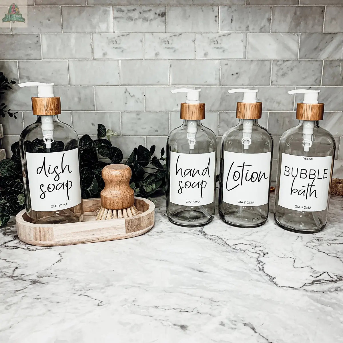 Four 16oz Glass Clear | White Label bottles—labeled for dish soap, hand soap, lotion, and bubble bath—are displayed on a marble counter with a scrub brush, complementing a light gray tiled wall and nearby greenery.
