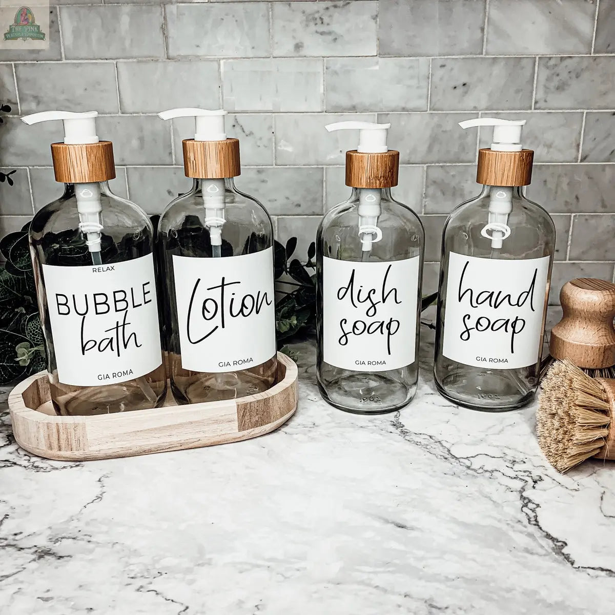 Four 16oz Glass Clear bottles with white waterproof labels—bubble bath, lotion, dish soap, and hand soap—are neatly arranged on a bathroom counter, accompanied by a wooden dish and two scrub brushes in front of white tile.