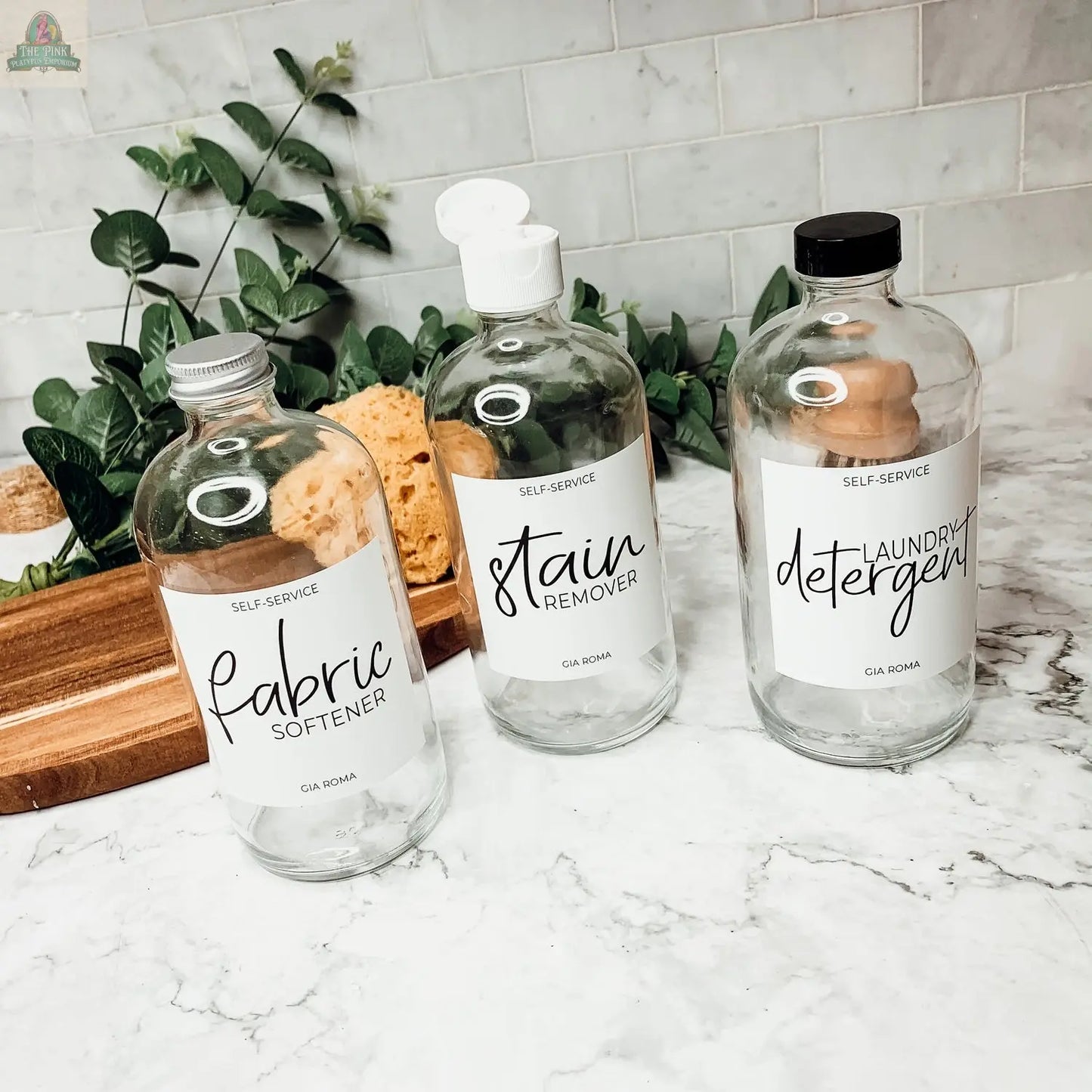 Three 16oz Glass Clear bottles with white labels for Fabric Softener, Stain Remover, and Laundry Detergent rest on a marble countertop with waterproof labeling. Green plants and sponges enhance the modern display.
