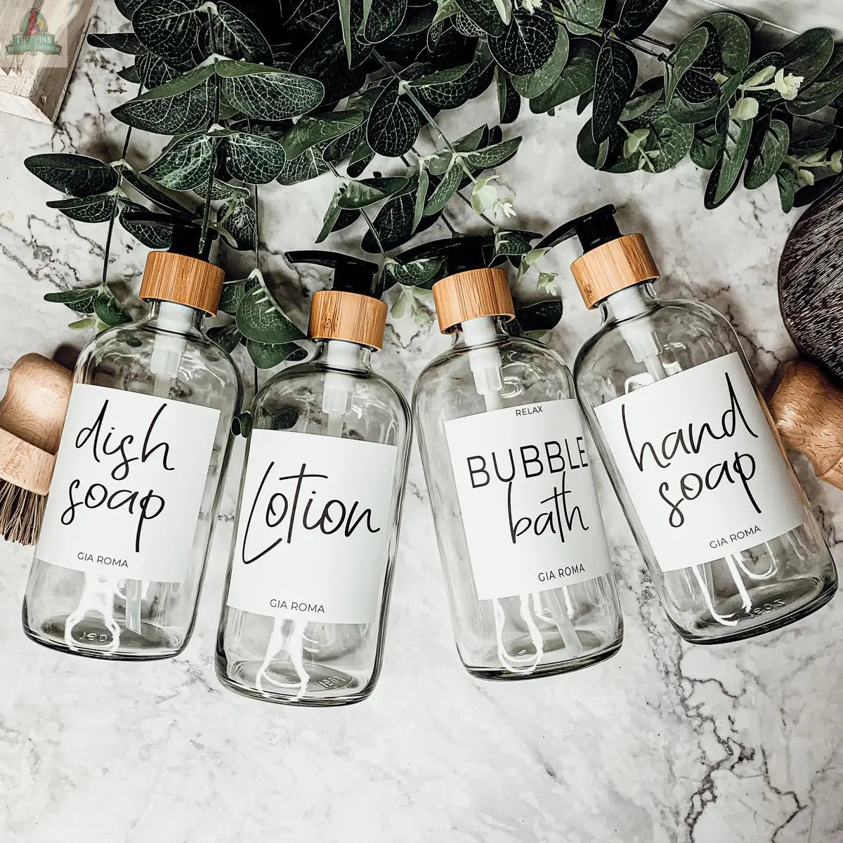 Four 16oz Glass Clear bottles with white labels, each filled with dish soap, lotion, bubble bath, and hand soap, are arranged on a marble surface with green leafy branches beside each elegant dispenser.