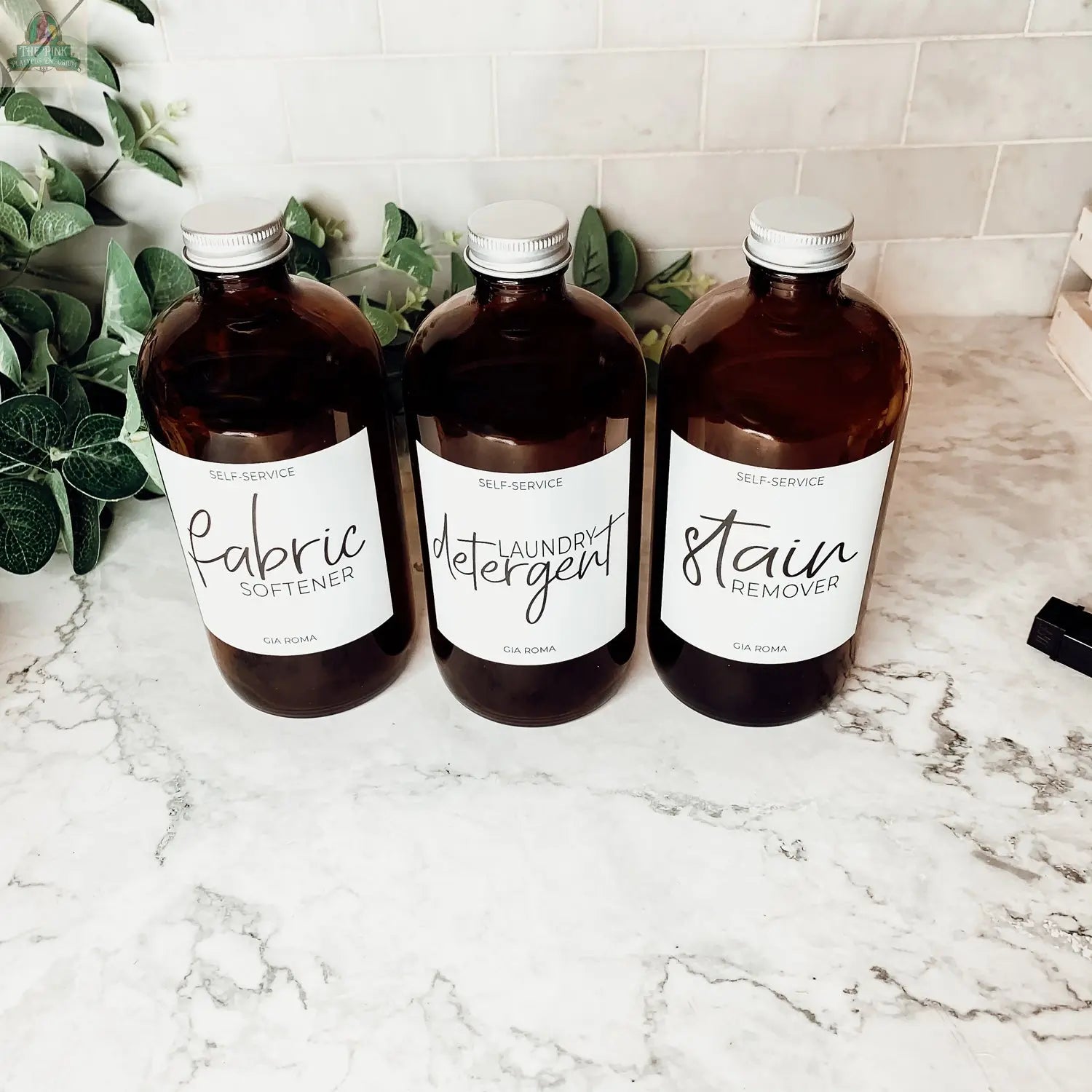 Three 16oz clear glass bottles with white labels for Fabric Softener, Laundry Detergent, and Stain Remover sit on a marble countertop with green plants behind them, each featuring a stylish waterproof design.