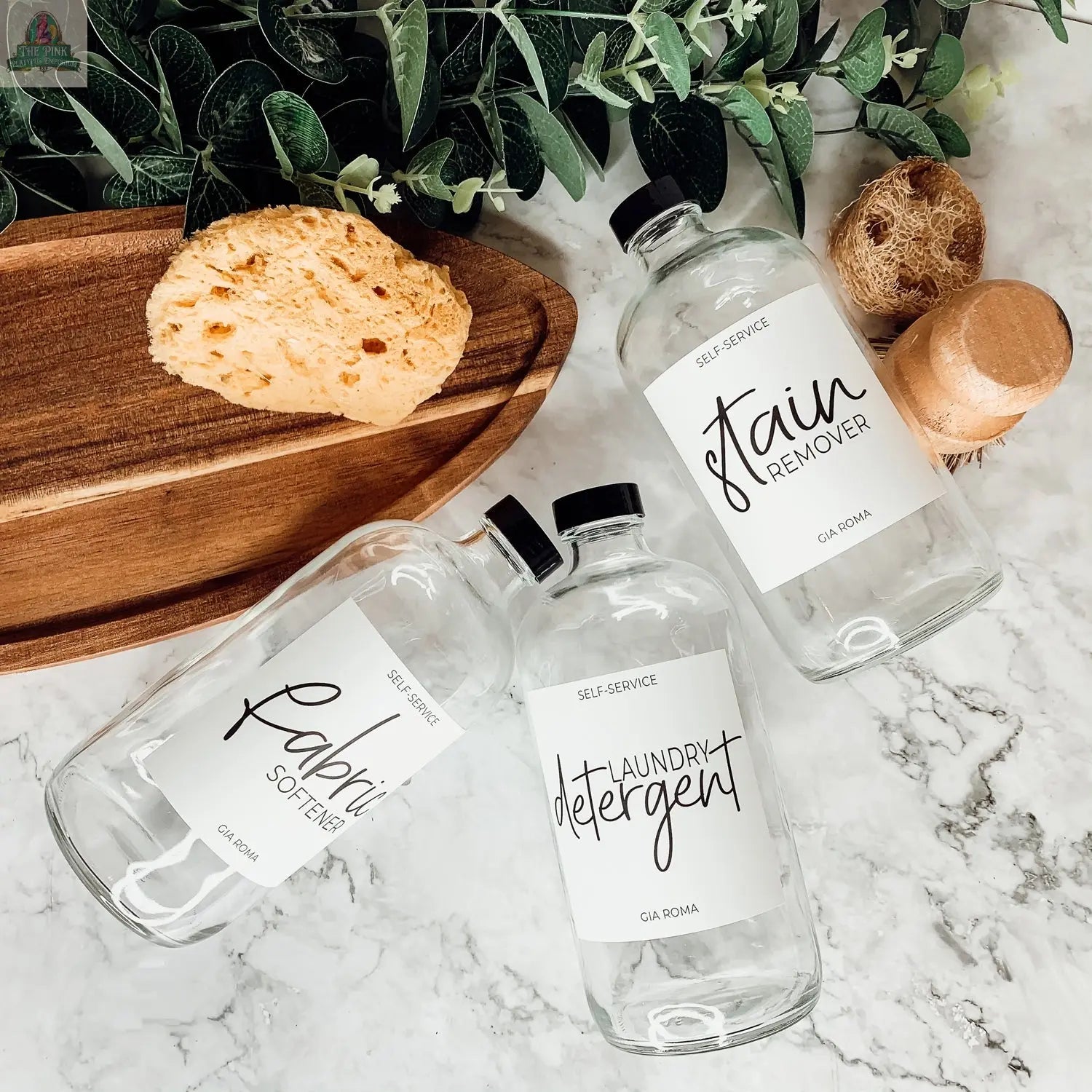 Three 16oz Glass Clear bottles with white labels—Stain Remover, Fabric Softener, and Laundry Detergent—rest on a marble surface alongside a clear glass dispenser, sponges, a wooden tray, a brush, and green plants.