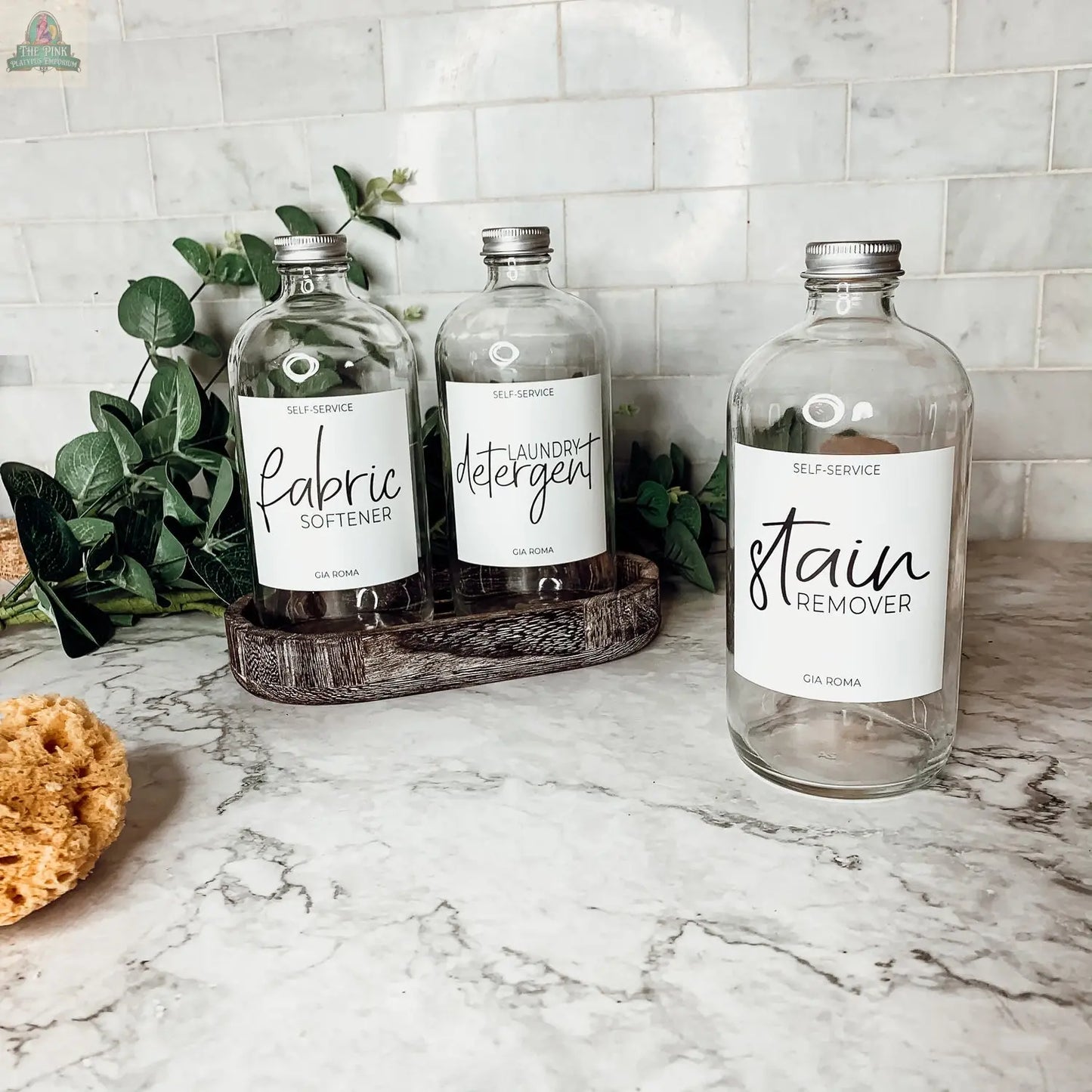 Three 16oz Glass Clear | White Label bottles labeled fabric softener, laundry detergent, and stain remover sit on a counter with a sponge and green plant, against a white tiled backsplash. Each glass dispenser features a stylish waterproof label.