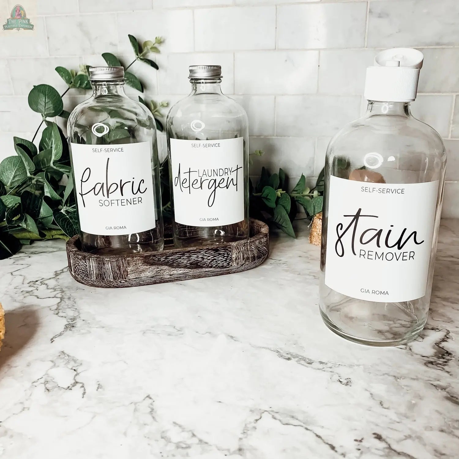 Three 16oz Glass Clear | White Label bottles—labeled for fabric softener, laundry detergent, and stain remover—sit on a marble countertop; two rest on a wooden tray with green foliage, while one stands alone.