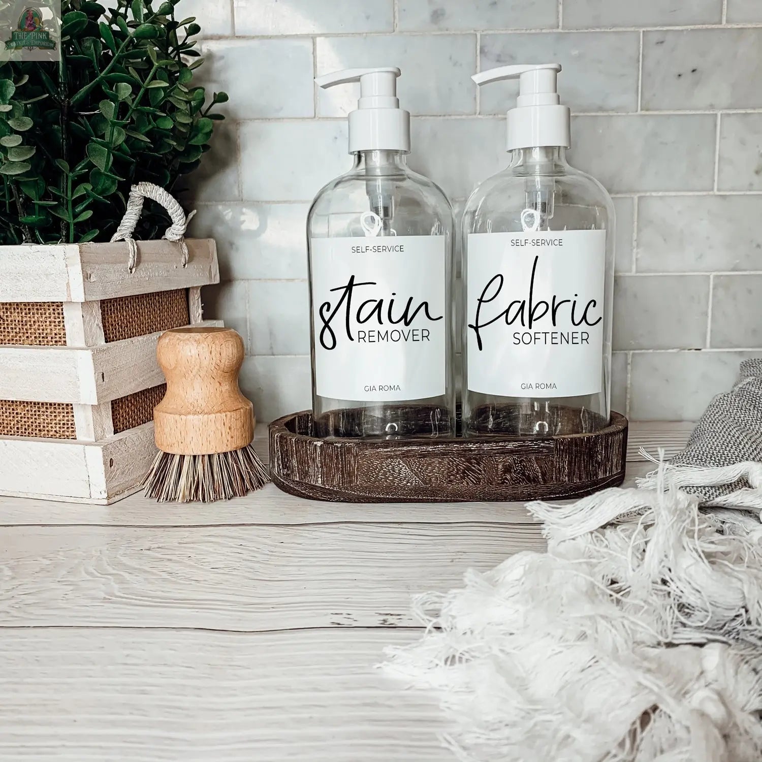Two 16oz Glass Clear | White Label dispenser bottles labeled Stain Remover and Fabric Softener sit on a wooden tray on a white countertop, featuring waterproof labels, with a scrub brush, potted plant, and white towels nearby.