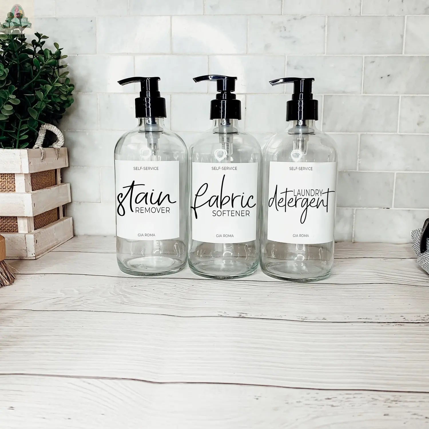 Three 16oz Glass Clear bottles with white labels—Stain Remover, Fabric Softener, and Laundry Detergent—are arranged on light wood with white tile and greenery, creating a clean, organized look.