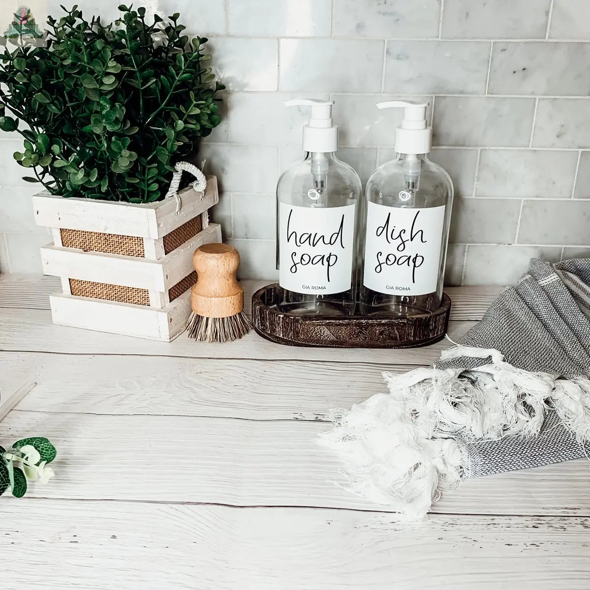 A kitchen counter features a potted plant, a wooden scrub brush, two 16oz Glass Clear | White Label dispensers (for hand and dish soap), and a gray striped towel with white fringe against a tiled backsplash.