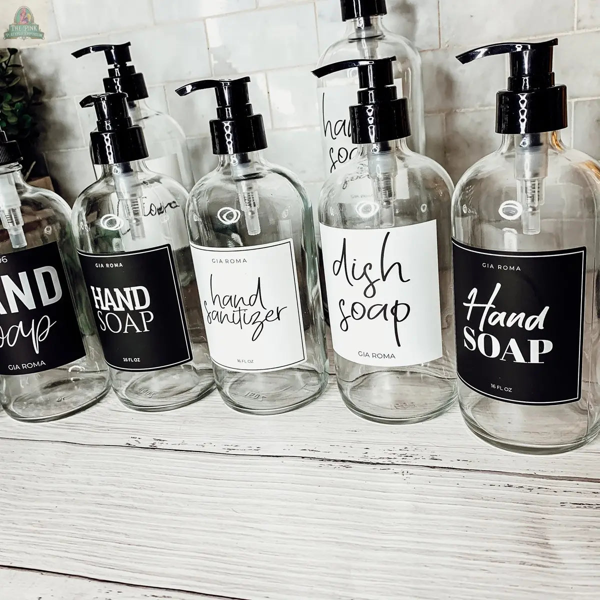 Six 16oz Glass Clear bottles with White Labels are arranged on a white wooden counter against a tiled backsplash. Each dispenser is boldly labeled in black and white for hand soap, sanitizer, and dish soap.
