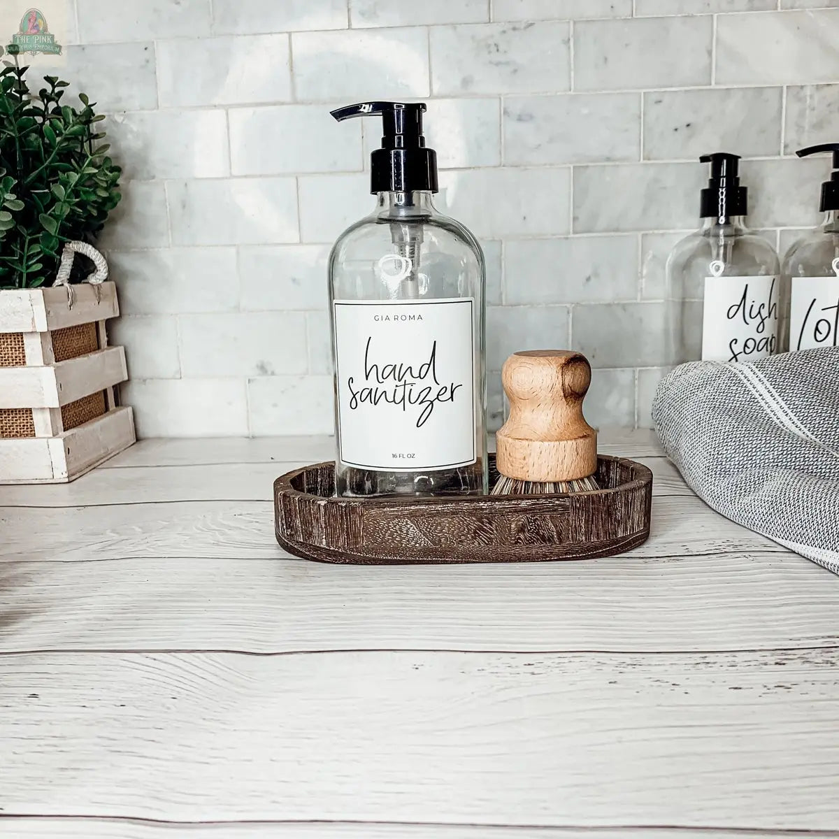 The 16oz Glass Clear | White Label hand sanitizer dispenser with a black pump sits on a wooden tray beside a round scrub brush, while other cleaning products, a plant, and cloth are in the background on a white countertop.