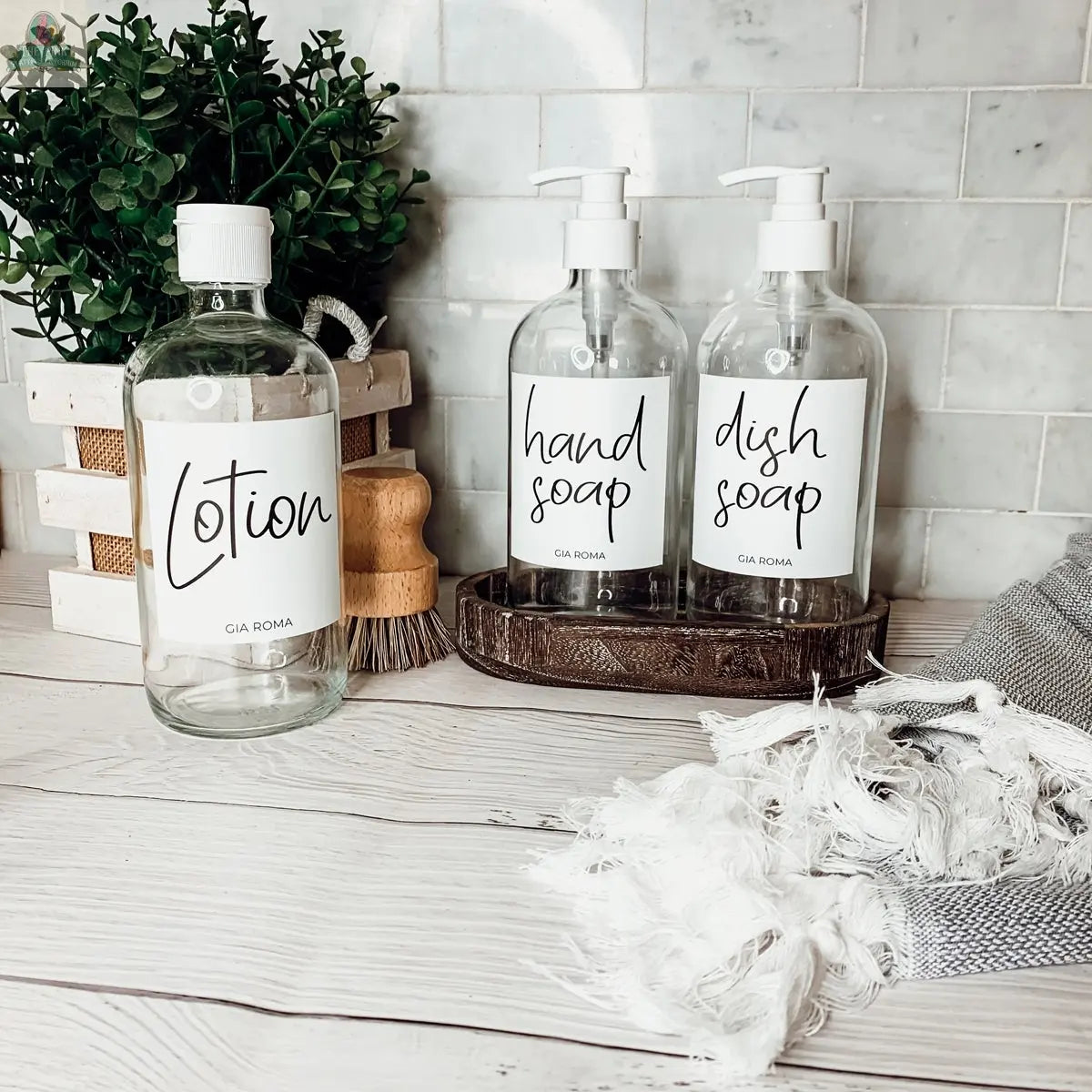 16oz Glass Clear bottles with white waterproof labels for “Lotion,” “hand soap,” and “dish soap” are on a white countertop with a plant, wooden brush, and fringed towel, set against white subway tiles.