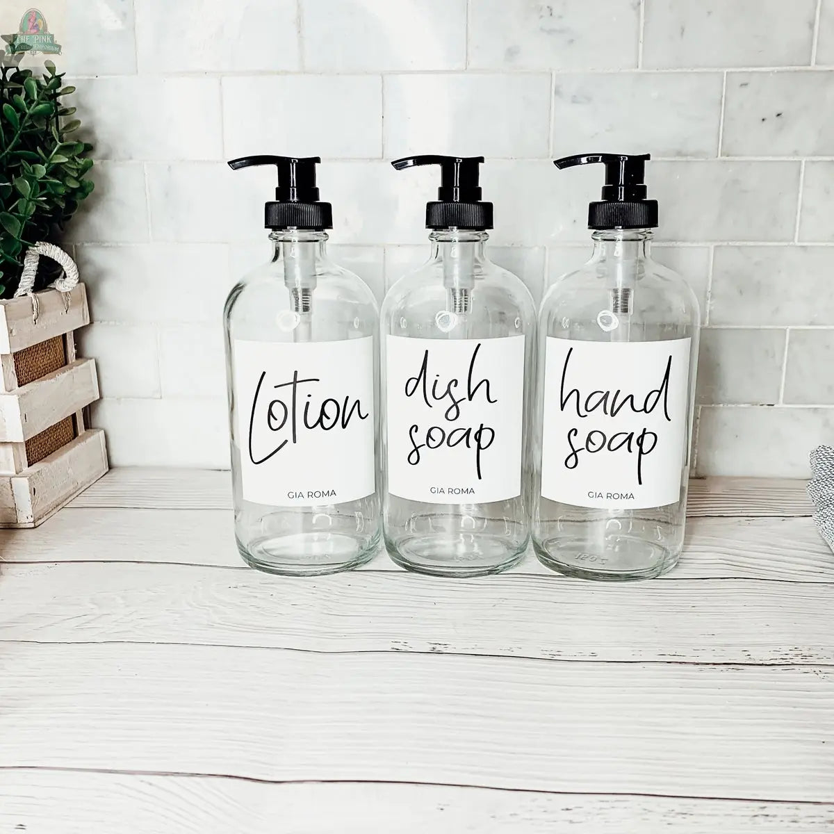 Three 16oz Glass Clear bottles with white waterproof labels—Lotion, Dish Soap, and Hand Soap—are displayed on a light wooden countertop before a white tiled backsplash, accented by a small plant and woven basket to the left.