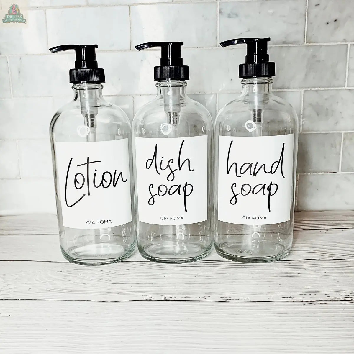 Three 16oz Glass Clear bottles with White Labels for Lotion, Dish Soap, and Hand Soap are arranged side by side on a white wooden surface against a white tiled background.