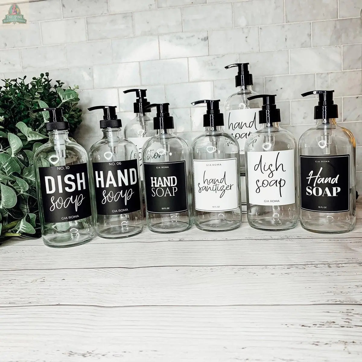 Eight 16oz Glass Clear | White Label bottles with “Hand Soap,” “Dish Soap,” and “Hand Sanitizer” labels sit on a white wooden surface beside a plant, against a white tiled wall.