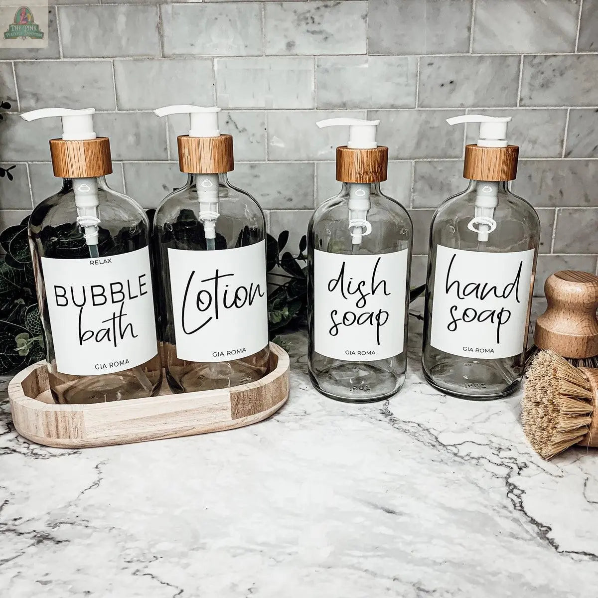 Four 16oz Glass Clear | White Label bottles, labeled for bubble bath, lotion, dish soap, and hand soap, sit on a marble counter with a tile backsplash, alongside wooden brushes.