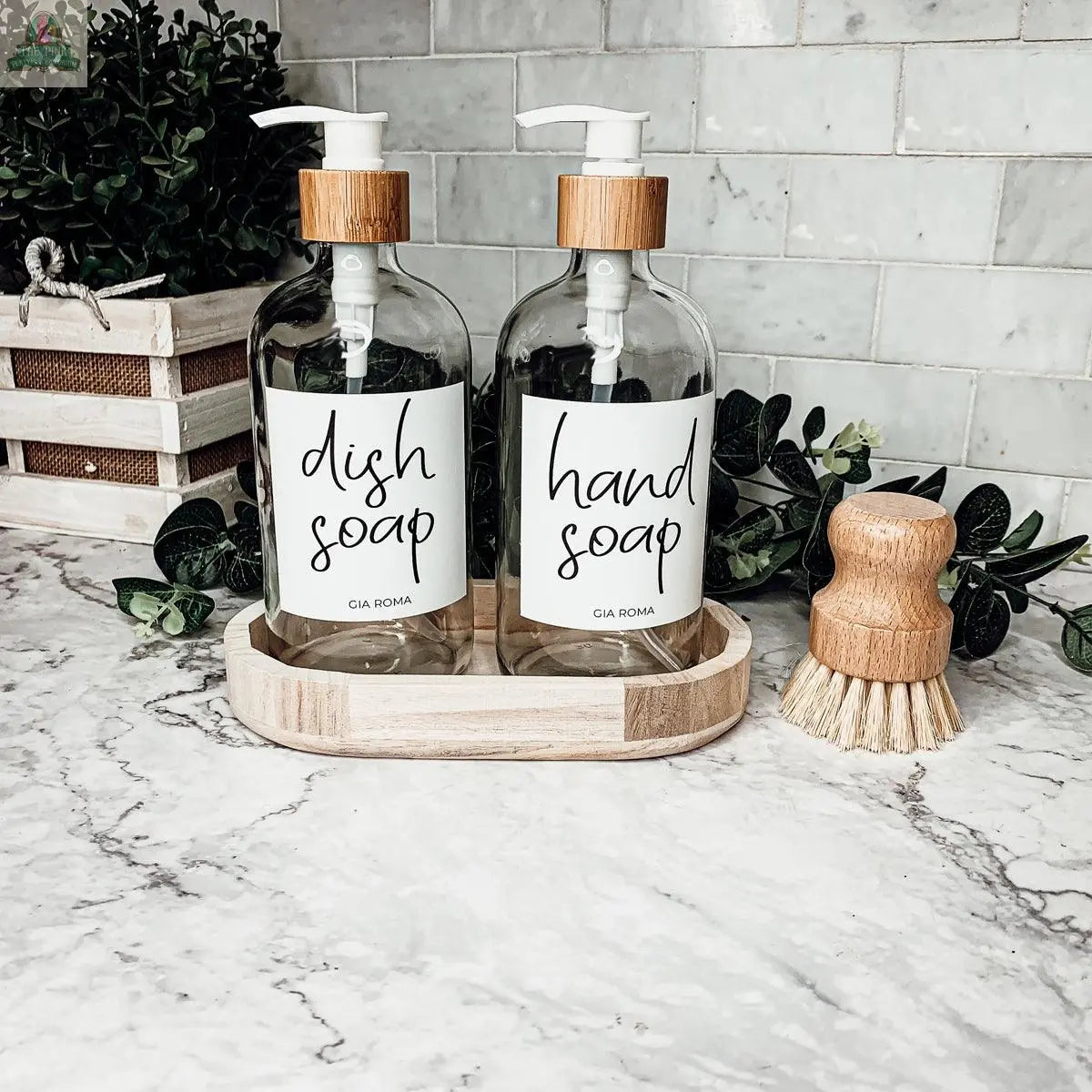 Two 16oz Glass Clear bottles with white labels for dish and hand soap are displayed on a wooden tray atop a marble countertop, along with a scrub brush, green plants, and a white basket in the background.