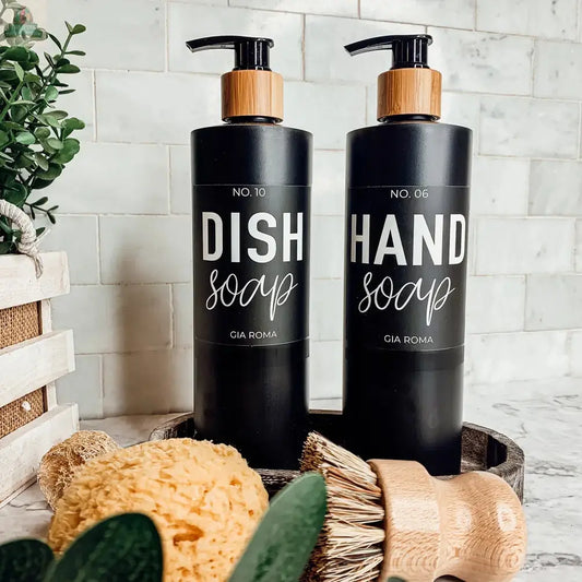 Two 16oz Plastic Black Bottles labeled Dish Soap and Hand Soap sit on a tray with a yellow sponge, wooden brush, and green leaves, set against white tiles. Each refillable bottle features a sleek, waterproof label.