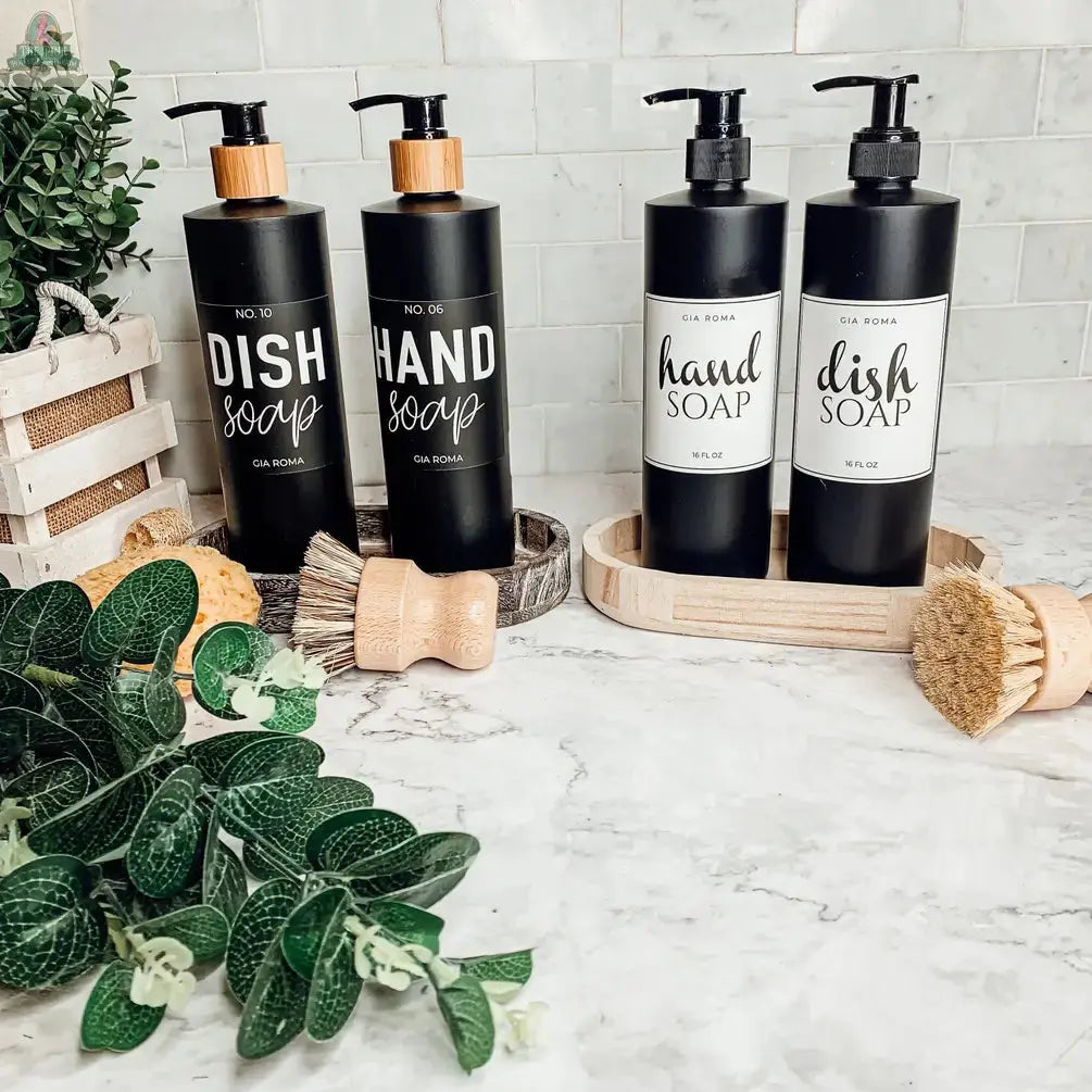 Four 16oz Plastic Black Bottles, labeled for dish soap and hand soap, rest on trays atop a marble counter with green plants, wooden brushes, sponges, and a white tiled backsplash. Each refillable dispenser has a clear waterproof label.