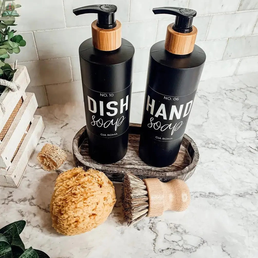 Two 16oz Plastic Black Bottles labeled Dish Soap and Hand Soap with waterproof labels and bamboo pumps sit on a tray beside a yellow sponge, wooden scrub brush, loofah, and green plants on a marble countertop. Refillable dispenser design.