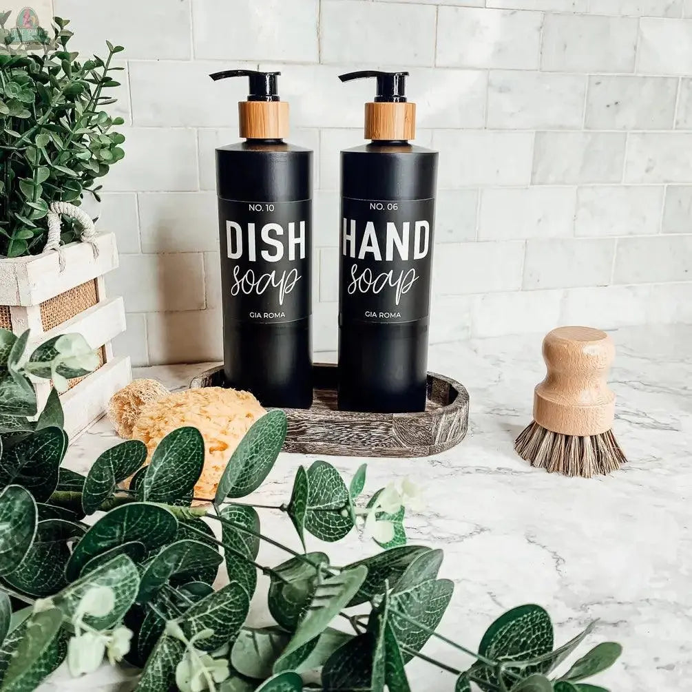 Two 16oz Plastic Black Bottles labeled Dish Soap and Hand Soap rest on a tray atop a marble counter with green plants, a scrub brush, and yellow sponge nearby. Each refillable dispenser has a waterproof label; a tiled backsplash is behind them.