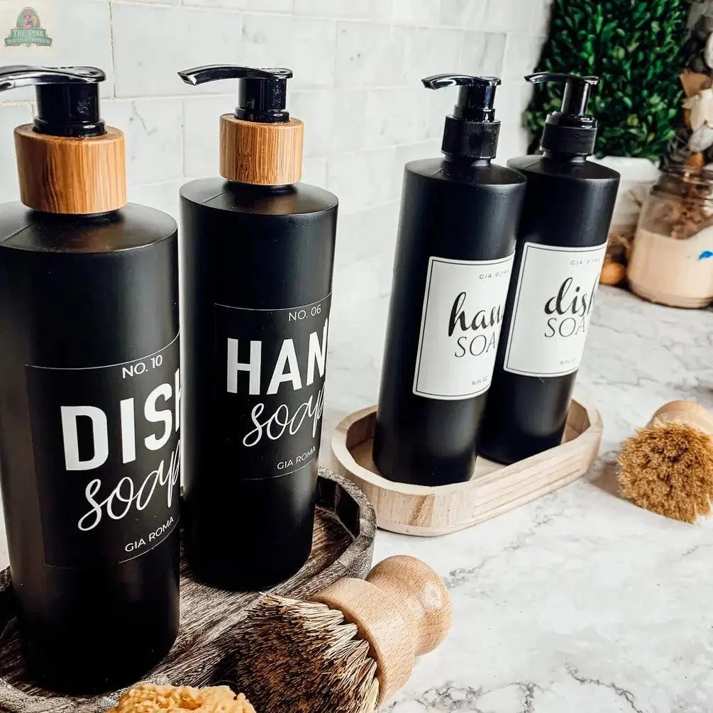 Four 16oz Plastic Black Bottles with waterproof Dish Soap and Hand Soap labels are displayed in wooden trays on a marble countertop, paired with scrub brushes, standing out against a plant and modern decor.