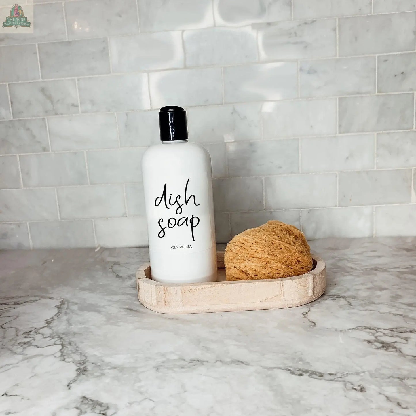 A 16oz Plastic Black Bottle with a waterproof label and a natural sponge rest on a light wooden tray atop a marble countertop, with white subway tile backsplash in the background.