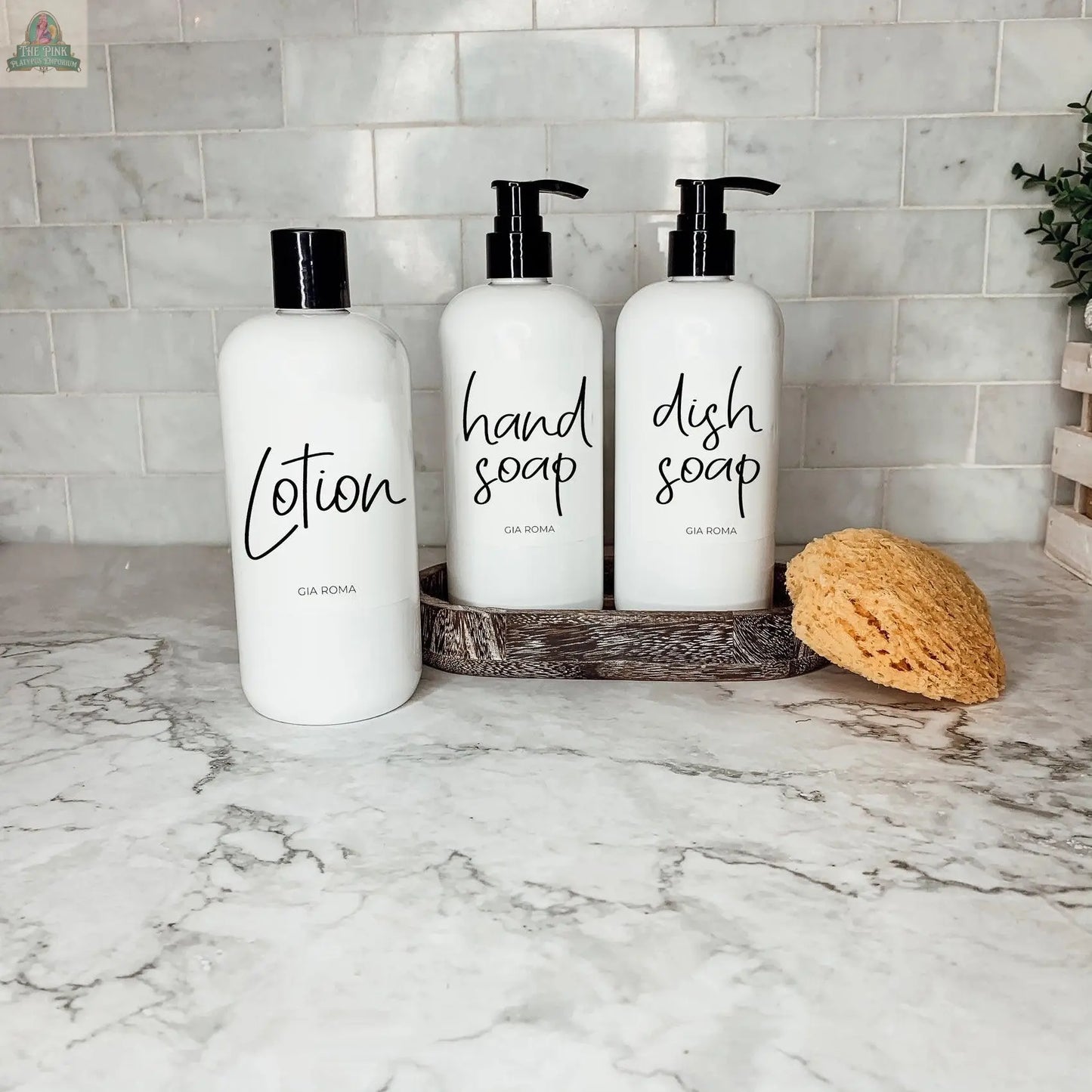 Three 16oz Plastic Black Bottles labeled lotion, hand soap, and dish soap sit on a counter with a yellow sponge by white tiles. Their waterproof labels keep them neat and organized in a small tray.