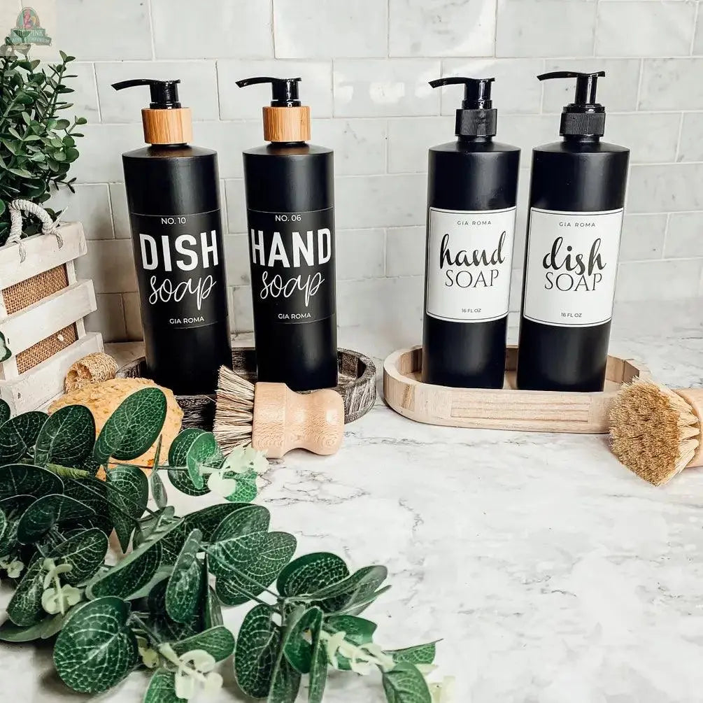 Four 16oz Plastic Black Bottles labeled “dish soap” and “hand soap” with waterproof labels rest on a marble counter, surrounded by green plants, wooden brushes, and a white tile backsplash.