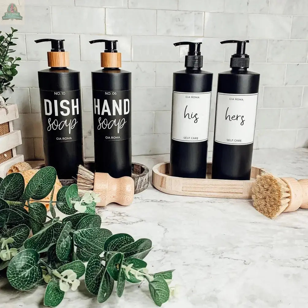 Four 16oz Plastic Black Bottles labeled Dish Soap, Hand Soap, His, and Hers are displayed on a marble counter with a scrub brush, greenery, and waterproof labels in front of a white tile backsplash.