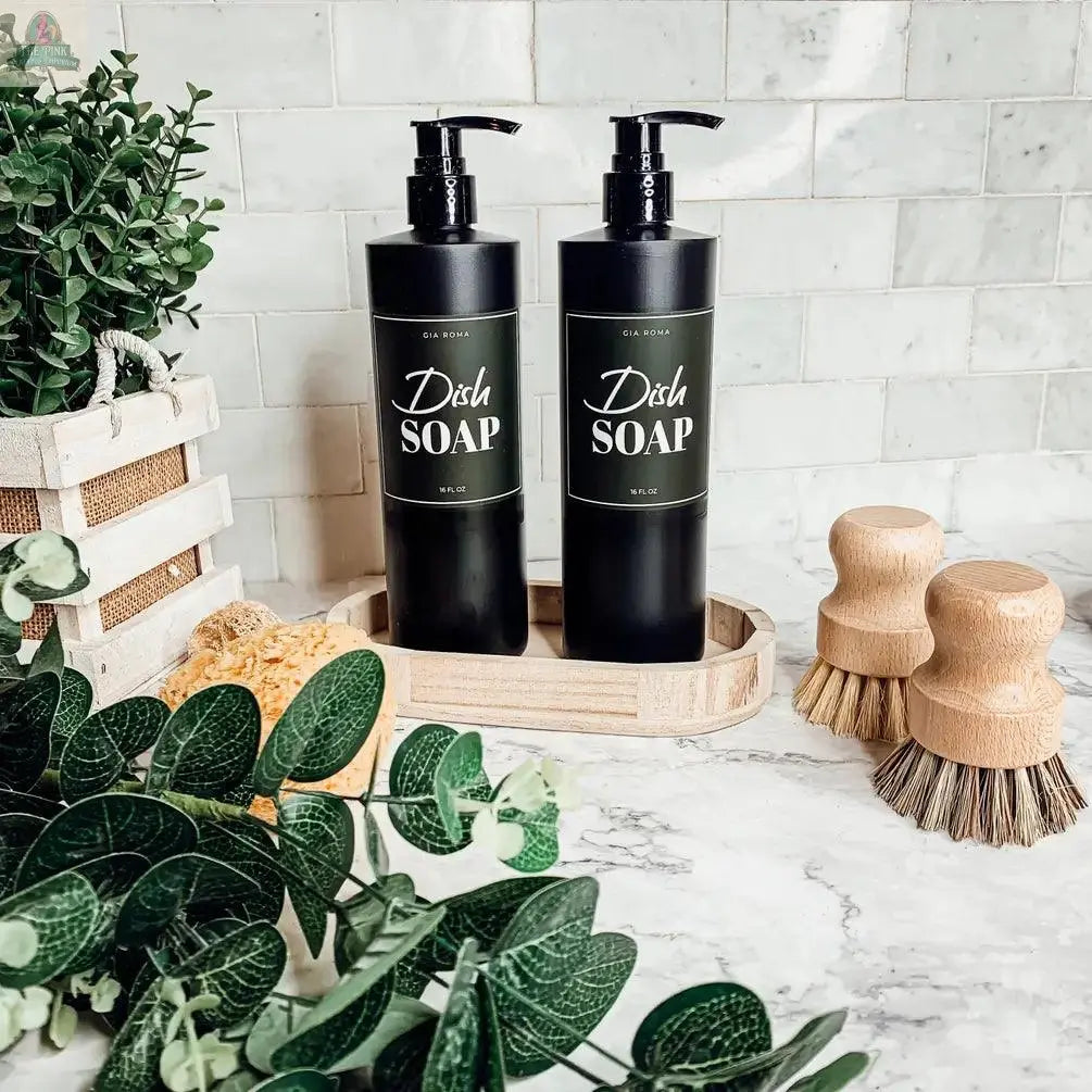 Two 16oz Plastic Black Bottles labeled Dish Soap sit on a wooden tray atop a marble countertop, surrounded by green plants and wooden dish brushes, with a tiled backsplash in the background—sleek, refillable kitchen dispensers.