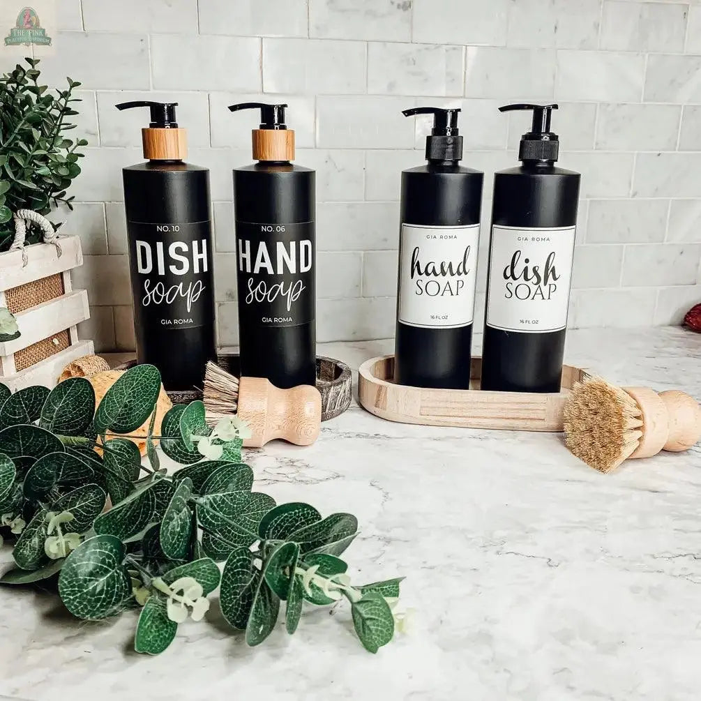 Four 16oz Plastic Black Bottles with waterproof labels for Dish Soap and Hand Soap are arranged on a marble countertop, surrounded by green leaves, a wooden brush, and a white tile backsplash.