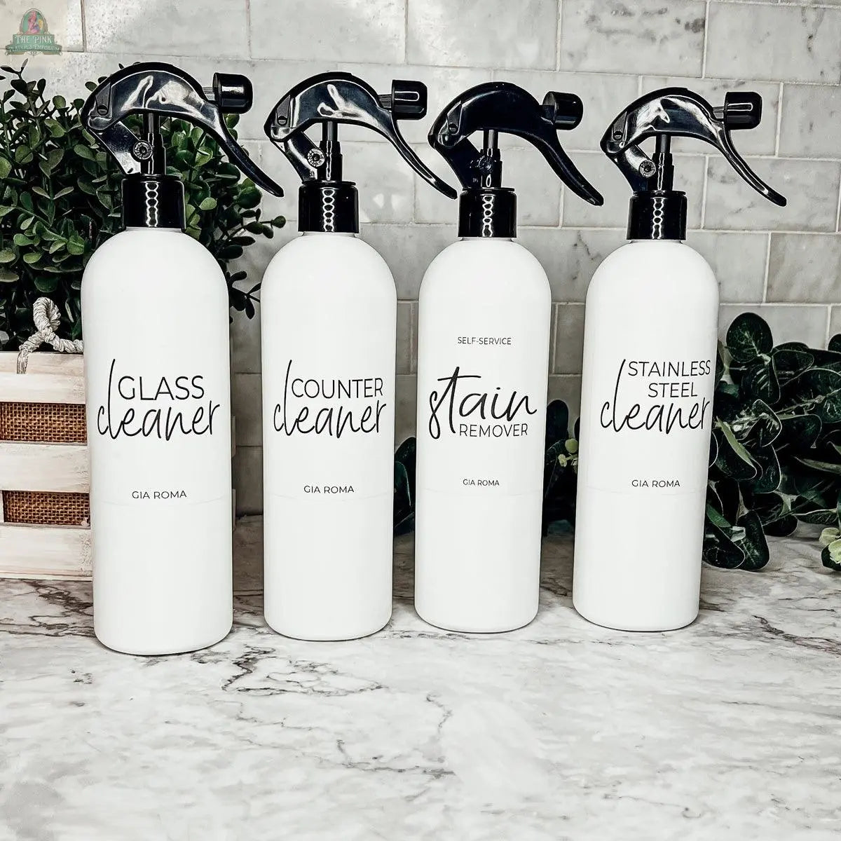 Four 16oz Plastic Cleaning Bottles—Glass Cleaner, Counter Cleaner, Stain Remover, Stainless Steel Cleaner—stand on a marble countertop with green plants and white tile in the background. Reusable dispensers for every need.