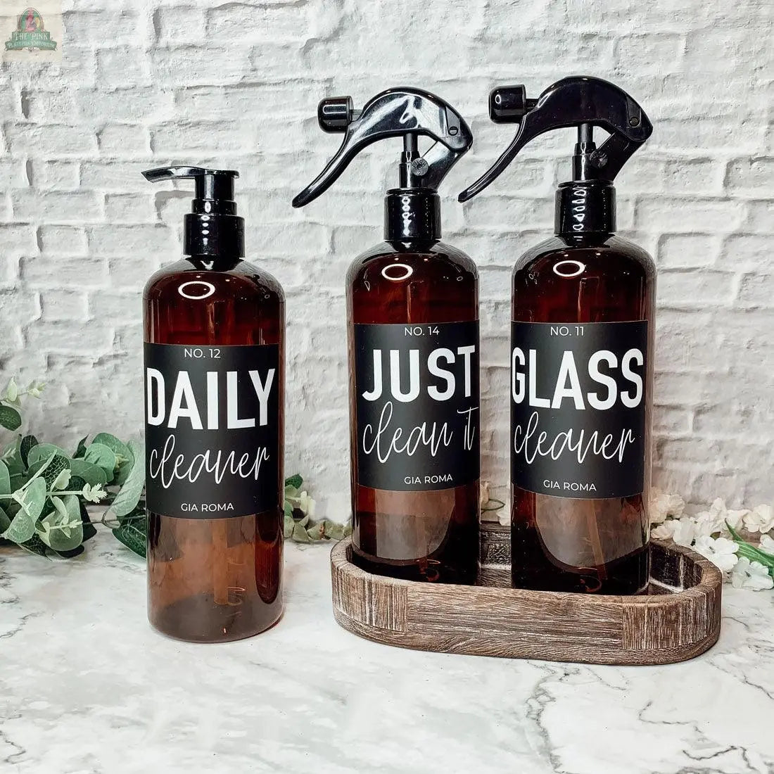 Three 16oz Plastic Cleaning Bottles with waterproof labels—DAILY Cleaner, JUST clean it, and GLASS Cleaner—sit on a marble counter with a tray and green foliage, offering stylish, reusable dispensers for any space.
