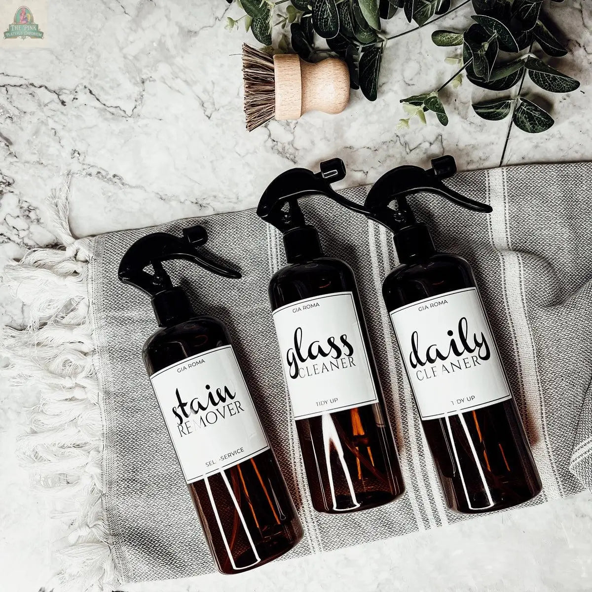 Three 16oz Plastic Cleaning Bottles with waterproof labels—stain remover, glass cleaner, and daily cleaner—are displayed on a gray towel with a scrub brush and green leaves. Ideal reusable dispensers for your cleaning routine.