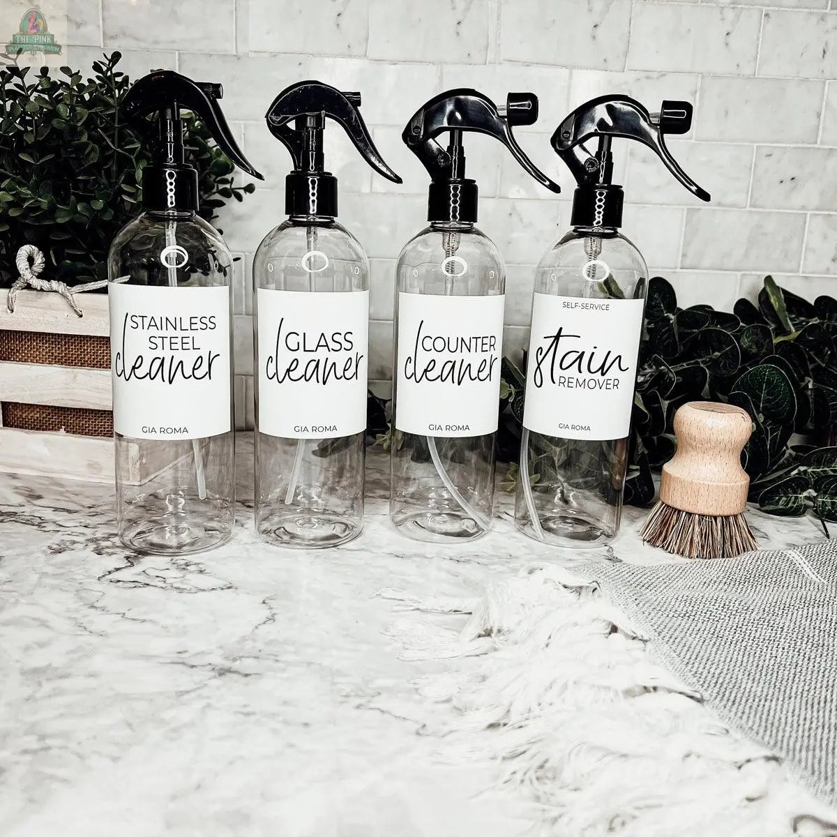 Four 16oz Plastic Cleaning Bottles, each labeled for stainless steel cleaner, glass cleaner, counter cleaner, and stain remover, are lined up on a marble countertop with green plants and a scrubbing brush in the background.