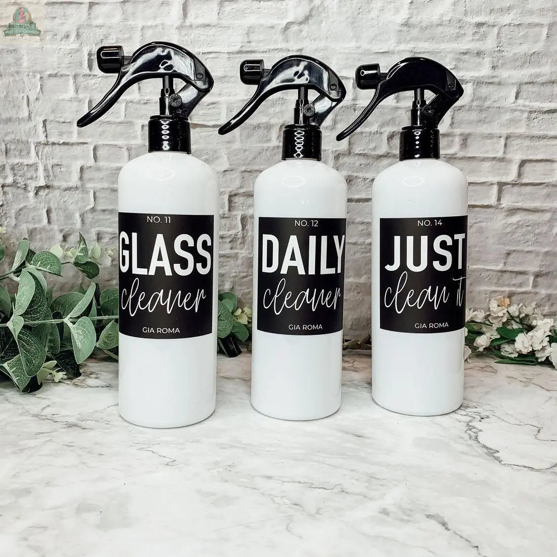 Three 16oz Plastic Cleaning Bottles with black waterproof labels—“GLASS cleaner,” “DAILY cleaner,” and “JUST clean it”—stand side by side on a marble surface, with a white brick wall and greenery in the background.