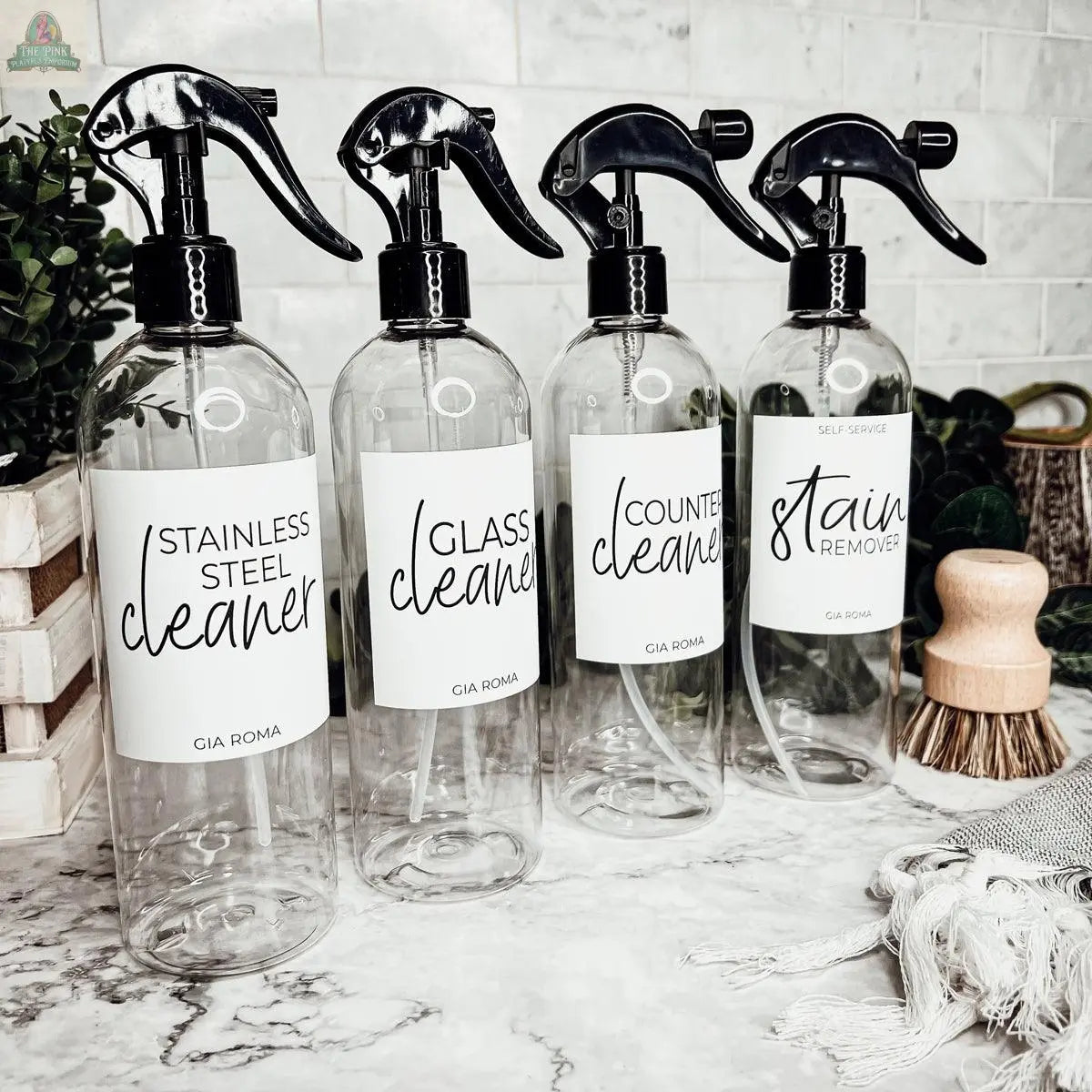 Four 16oz Plastic Cleaning Bottles—labeled for stainless steel, glass, counter, and stain remover—are arranged on a marble countertop with reusable dispensers, cleaning brushes, and green plants.