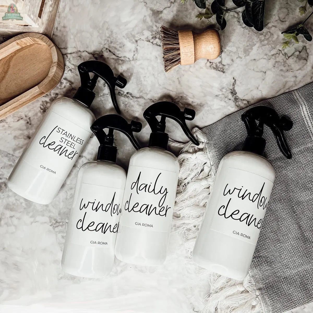 Four 16oz Plastic Cleaning Bottles with waterproof labels—stainless steel cleaner, daily cleaner, and window cleaner—are arranged on a marble countertop beside a tray, brush, and gray towel as stylish reusable dispensers.