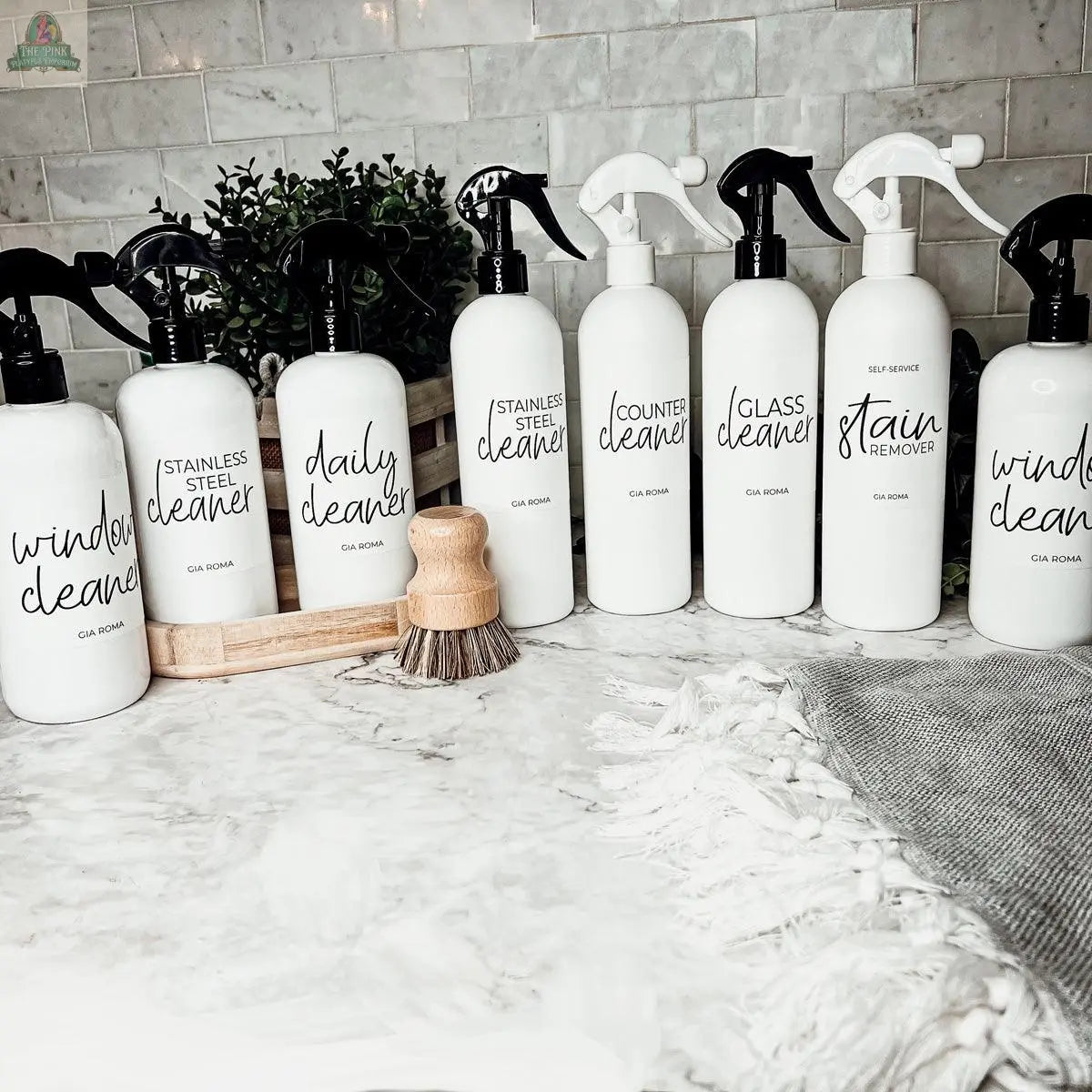 Eight 16oz Plastic Cleaning Bottles, each with waterproof labels, are arranged on a countertop near a scrub brush, gray towel, and potted plant—creating a clean and organized display.