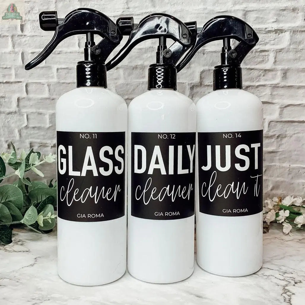 Three 16oz Plastic Cleaning Bottles—labeled GLASS cleaner, DAILY cleaner, and JUST clean it—are arranged side by side on a marble surface with green plants and a white brick wall in the background.