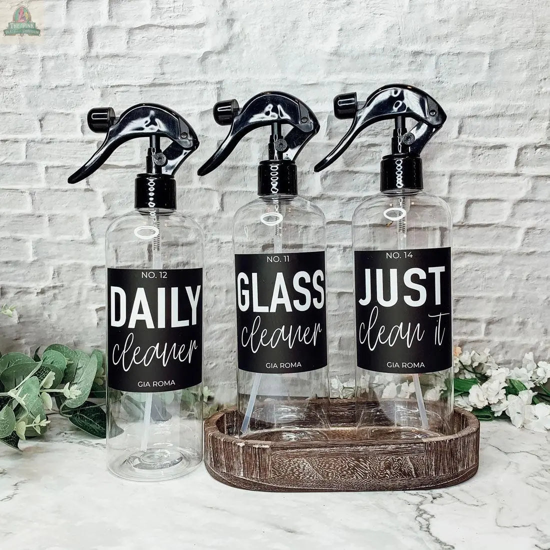 Three 16oz Plastic Cleaning Bottles labeled DAILY cleaner, GLASS cleaner, and JUST clean it rest on a wooden tray by a white brick wall, accented by greenery. These reusable bottles add a stylish touch to your cleaning routine.